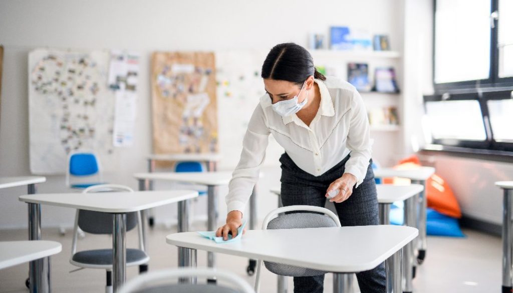 teacher wearing mask in a classroom disinfecting desks