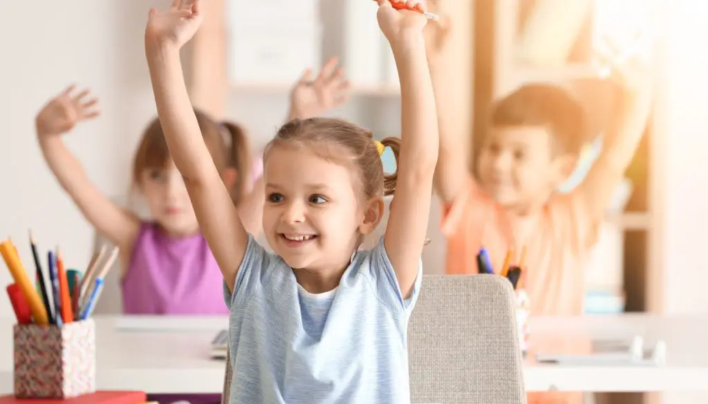 kids sitting at desks in classroom with hands up smiling