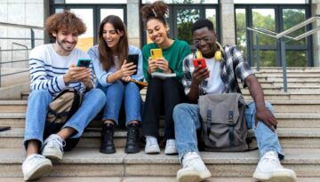 Multiracial college student friends look mobile phone laughing together. University students using smartphone outdoors.